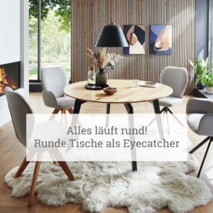 Ein modernes Esszimmer mit einem runden Holztisch in der Mitte, umgeben von vier Stühlen mit einer Kombination aus weißen und hölzernen Elementen. Über dem Tisch hängt eine große schwarze Pendelleuchte. Links im Raum befindet sich ein Kamin mit sichtbaren Flammen hinter Glas. Der Boden ist mit einem großen, strukturierten weißen Fellteppich bedeckt. Im Hintergrund gibt es ein großes Fenster mit Blick auf das Grün draußen, und die Wand rechts ist mit vertikalen Holzpaneelen und zwei abstrakten Wandkunstwerken in Erdtönen verziert. Der Text im Bild lautet: ‘Alles läuft rund! Runde Tische als Eyecatcher.’