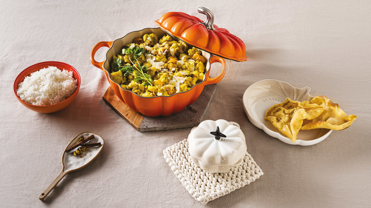 Herbstliches Tisch-Setting mit einem orangefarbenen Kürbisbräter, gefüllt mit Curry und frischen Kräutern, dazu Schälchen mit Reis, knusprigen Chips und Gewürzen – stilvoll angerichtet auf hellem Tischtuch.“